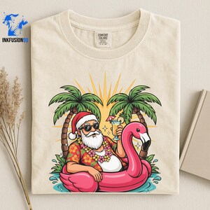 May include: A cream-colored t-shirt featuring a graphic of Santa Claus wearing sunglasses and a floral shirt, sitting in a pink flamingo pool float between two palm trees. Santa holds a tropical drink, and a flower lei hangs around the neck.