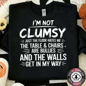 Peut inclure: Sweat-shirt noir avec texte blanc : « I'M NOT CLUMSY JUST THE FLOOR HATES ME THE TABLE & CHAIRS ARE BULLIES AND THE WALLS GET IN MY WAY ». Un logo de téléchargement numérique est dans le coin inférieur droit.