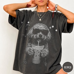 May include: Dark gray t-shirt featuring a black and white graphic of a dog wearing sunglasses and holding a beverage with a straw. The dog is the central focus, with a blurred background. The shirt has a relaxed fit.