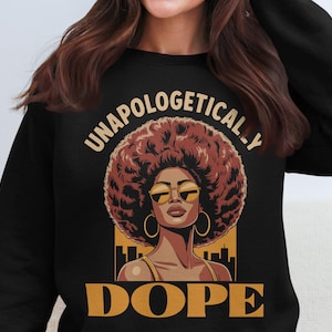 Puede incluir: Sudadera negra con un gráfico de una mujer con afro, gafas de sol y aros dorados. Las palabras "UNAPOLOGETICALLY DOPE" están impresas encima y debajo de la imagen en letras doradas. La mujer está situada contra un paisaje urbano amarillo y negro.