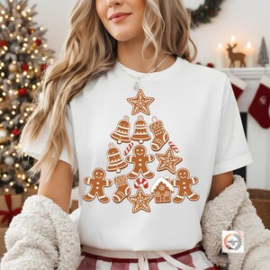 Könnte beinhalten: Weißes T-Shirt mit einem Weihnachtsbaum-Design aus Lebkuchen. Der Baum besteht aus Lebkuchenmännern, Sternen, Glocken, einem Haus und einem Strumpf. Die Kekse sind braun mit weißem Zuckerguss und roten Akzenten.