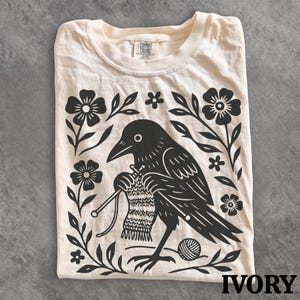 May include: An ivory t-shirt featuring a black graphic print of a crow knitting with two needles and a ball of yarn. The bird is surrounded by a decorative border of flowers and leaves. The inner neck label displays the text Comfort Colors.