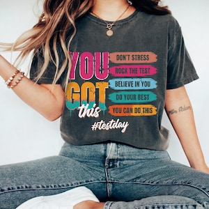 May include: A dark gray t-shirt with the words "YOU GOT this" in pink, orange, and blue. The shirt also has the phrases "DON'T STRESS", "ROCK THE TEST", "BELIEVE IN YOU", "DO YOUR BEST", and "YOU CAN DO THIS". The hashtag "#testday" is also visible.