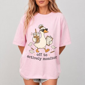 May include: Pink t-shirt with a cartoon goose wearing sunglasses, holding a coffee and clipboard. The text "off to actively monitor" is below the goose. The shirt is a light pink color.