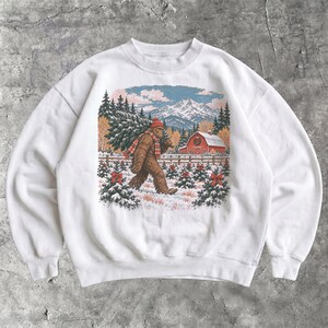 May include: A white crewneck sweatshirt featuring a graphic print of a brown Bigfoot creature wearing a red beanie and a red and white plaid scarf, carrying a pine tree through a snowy landscape with a red barn and mountains.