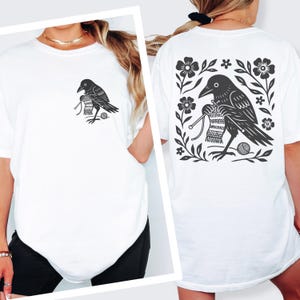 May include: A white t-shirt featuring a black graphic print of a crow knitting with needles and yarn. The front shows a small version of the crow on the chest, while the back displays a larger version of the same crow surrounded by floral motifs.