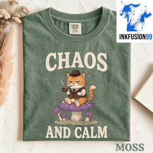 May include: A moss green t-shirt featuring a graphic of an orange tabby cat wearing a brown bowler hat and vest while smoking a pipe. The cat sits on a large purple mushroom. The text "CHAOS AND CALM" is printed in white above and below the cat.