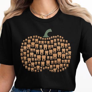 Peut inclure: T-shirt noir avec un motif de citrouille fait de dents. La citrouille est composée de dents de différentes tailles et formes dans des tons bruns. Une tige verte surmonte la citrouille.