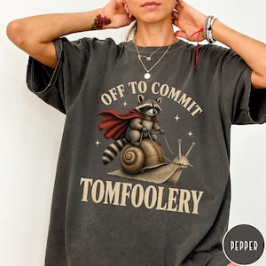 May include: A charcoal gray t-shirt featuring a graphic of a raccoon wearing a red cape and riding a snail. The text above reads OFF TO COMMIT and the text below reads TOMFOOLERY. The design includes small star accents around the central figures.