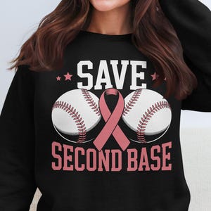 Peut inclure: Sweat-shirt noir avec l'inscription "SAVE SECOND BASE" en blanc et rose. Deux balles de baseball avec des coutures rouges et un ruban rose sont au centre. Deux étoiles roses sont au-dessus du mot "SAVE".
