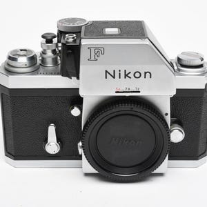 May include: A silver and black Nikon F 35mm film camera body with a black lens cap. The camera features a silver pentaprism housing with the letter F and the Nikon logo printed on the front, along with various dials, levers, and buttons.