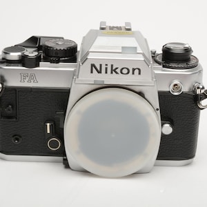 May include: Silver and black Nikon FA 35mm film camera with a lens cap. The camera body has the word "Nikon" printed on the front. The camera has various dials and buttons. The camera is a vintage photography item.