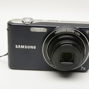 May include: A black Samsung digital camera with a silver top edge, a prominent Samsung Zoom Lens, and a 7x optical zoom indicator. The device features a built-in flash, a shutter button, and a small black wrist strap attached to the side of the body.