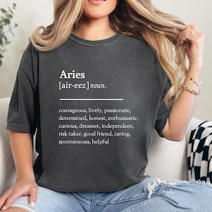 Könnte beinhalten: Ein graues T-Shirt mit dem Wort "Aries" in weißer Schrift, gefolgt von der phonetischen Aussprache in Klammern und dem Wort "Substantiv." Der Text setzt sich mit einer Liste von Persönlichkeitsmerkmalen fort, die mit dem Sternzeichen Widder verbunden sind.