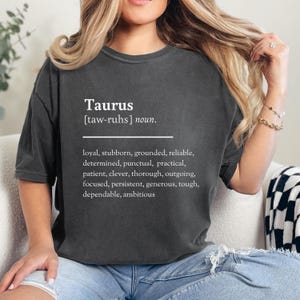 Op de afbeelding: Een donkergrijs T-shirt met het woord "Taurus" en de definitie in wit gedrukt. De definitie bevat de uitspraak "[taw-ruhs] noun." en een lijst met persoonlijkheidskenmerken: loyaal, koppig, nuchter, betrouwbaar, vastberaden, punctueel, praktisch, geduldig, slim, grondig, uitgaand, gefocust, volhardend, genereus, taai, betrouwbaar, ambitieus.