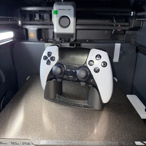 May include: A white and black video game controller resting on a black stand inside a 3D printer. The controller has a sleek design with various buttons and directional pads. The printer has the text "Bambu Lab" on the top.