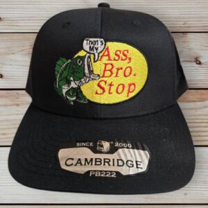 May include: Black baseball cap with a mesh back. The front features a patch with a cartoon fish and the text "That's My Ass, Bro. Stop" in a speech bubble. The hat also has a "Cambridge" label.