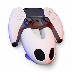 May include: A white and black PlayStation 5 controller on a white charging stand. The controller features black thumbsticks and buttons, with the PlayStation logo. The stand has a distinctive design with two large, black eye-shaped openings.
