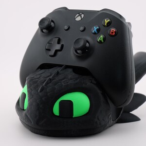 Pode incluir: Controlador Xbox preto apoiado em um suporte de controlador preto com tema de drag&atilde;o com olhos verdes brilhantes. O controlador tem bot&otilde;es coloridos e um D-pad. O suporte &eacute; texturizado e detalhado.
