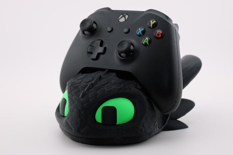 Pode incluir: Controlador Xbox preto apoiado em um suporte de controlador preto com tema de drag&atilde;o com olhos verdes brilhantes. O controlador tem bot&otilde;es coloridos e um D-pad. O suporte &eacute; texturizado e detalhado.