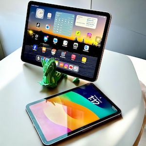 Op de afbeelding: Twee tablets met afgeronde hoeken en zwarte randen. Eén tablet wordt ondersteund door een felgroen, 3D-geprint drakenfiguurtje en toont een startscherm vol apps. De tweede tablet ligt plat en toont een kleurrijke, abstracte achtergrond.