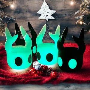 May include: Three glowing green and black figurines of a creature with large ears and a round head. The figurines are sitting on a red fuzzy surface with Christmas ornaments.