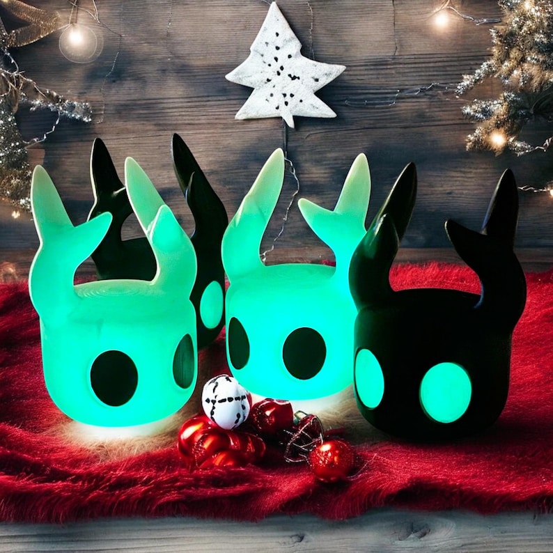 May include: Three glowing green and black figurines of a creature with large ears and a round head. The figurines are sitting on a red fuzzy surface with Christmas ornaments.