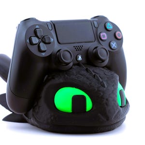 Pode incluir: Um controle preto do PlayStation 4 repousa sobre um suporte de controle preto com tema de drag&atilde;o. O suporte apresenta olhos verdes brilhantes. O controle tem o layout padr&atilde;o de bot&otilde;es com s&iacute;mbolos coloridos.