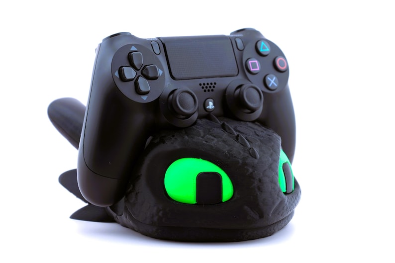 Pode incluir: Um controle preto do PlayStation 4 repousa sobre um suporte de controle preto com tema de drag&atilde;o. O suporte apresenta olhos verdes brilhantes. O controle tem o layout padr&atilde;o de bot&otilde;es com s&iacute;mbolos coloridos.