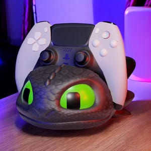 Pode incluir: Um controle PlayStation 5 preto e branco repousa sobre um suporte de carregamento preto com tema de drag&atilde;o. O drag&atilde;o tem olhos verdes brilhantes e um brilho vermelho. O controle tem al&ccedil;as brancas.