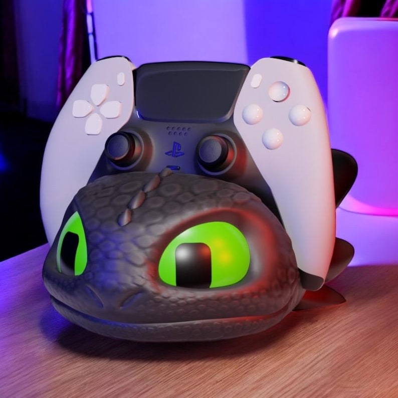 Pode incluir: Um controle PlayStation 5 preto e branco repousa sobre um suporte de carregamento preto com tema de drag&atilde;o. O drag&atilde;o tem olhos verdes brilhantes e um brilho vermelho. O controle tem al&ccedil;as brancas.