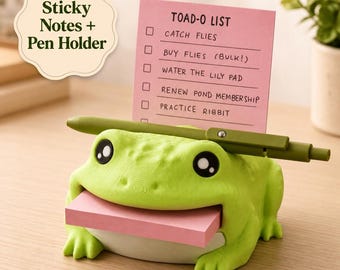May include: A light green frog-shaped desk organiser featuring a slot for a stack of pink sticky notes in its mouth and a horizontal olive green pen holder across its back. A pink notepad stands upright with the text TOAD-O LIST and tasks.