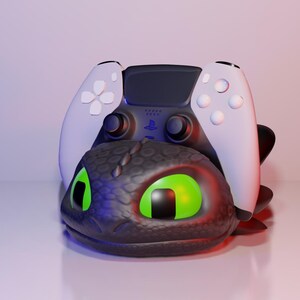 Pode incluir: Uma cabe&ccedil;a de drag&atilde;o preta e cinza com olhos verdes brilhantes suporta um controle PlayStation 5 branco e preto. O controle tem bot&otilde;es brancos e uma se&ccedil;&atilde;o central preta. A cabe&ccedil;a do drag&atilde;o tem uma superf&iacute;cie texturizada.