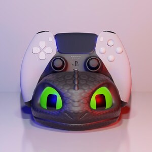 Pode incluir: Um suporte para comando PlayStation 5 com tema de drag&atilde;o preto e cinza. O suporte apresenta um design de cabe&ccedil;a de drag&atilde;o com olhos verdes brilhantes e um comando branco. O comando tem bot&otilde;es brancos e um centro preto.