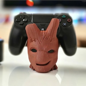 May include: A brown, wooden, Groot-inspired stand with a smiling face holds a black PlayStation 4 controller.