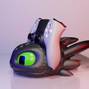 Pode incluir: Escultura estilizada de uma cabe&ccedil;a de drag&atilde;o preta com um olho verde e um controle de jogo preto e branco em cima. O drag&atilde;o tem reflexos vermelhos. O conjunto est&aacute; sobre uma superf&iacute;cie branca.