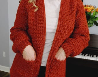 May include: A rust-colored crocheted cardigan with pockets. The cardigan is open in the front and has long sleeves.