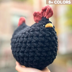May include: A handmade, black crochet chicken with red comb and wattle, and a yellow beak. The plush toy has a round body and is shown against a blurred background. The text "8+ COLORS" is in the upper right corner.