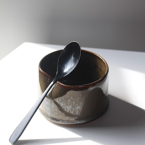May include: A ceramic bowl with a dark interior and a textured, light brown exterior, paired with a long, black spoon. The spoon rests on the bowl's rim, creating a minimalist aesthetic. The bowl is approximately 4 inches in diameter.