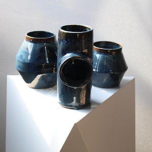 May include: Three ceramic vases in shades of blue with brown rims. The vases have unique shapes, including a T-shaped design. The vases are displayed on a white, geometric surface against a neutral background.