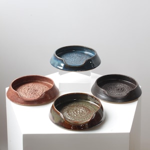 May include: Four circular ceramic grater plates with a textured center and a small spout on the rim. The plates are finished in glossy glazes of deep blue, reddish-brown, dark charcoal, and olive green, each featuring a raised pattern in the middle.