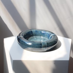 May include: A circular ceramic bowl with a thick, rounded rim and a deep center. The surface features a glossy glaze in shades of deep navy, cobalt, and lighter blue, with subtle vertical drip patterns and variations in color intensity throughout.