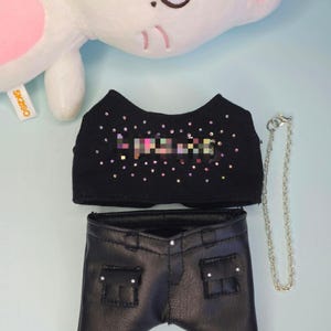 May include: A black doll outfit featuring a crop top and pants. The crop top is embellished with iridescent rhinestones. The pants are black faux leather with pockets. A silver chain is attached. A white plush toy with pink accents is in the background.