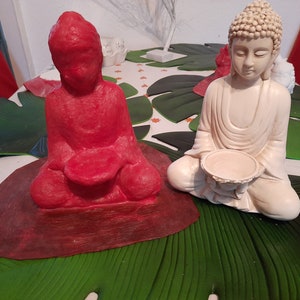 Könnte beinhalten: Zwei Buddha-Statuen, eine rote und eine weiße, sitzen auf grünen, blattartigen Matten. Beide Statuen halten eine kleine Schale in ihren Händen.