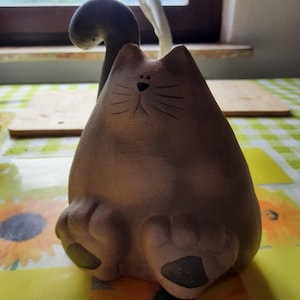 Puede incluir: Una figurita de cerámica de un gato de color marrón y blanco. El gato está sentado con la cola enroscada alrededor de su cuerpo. El gato tiene una cara pintada con ojos negros y bigotes.