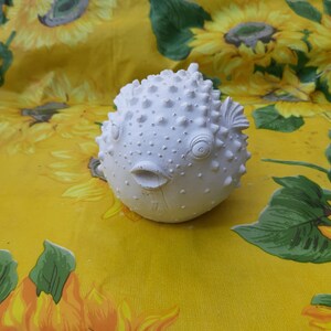 May include: White ceramic pufferfish figurine with a textured surface and a slightly open mouth. The figurine is sitting on a yellow background with a floral pattern.