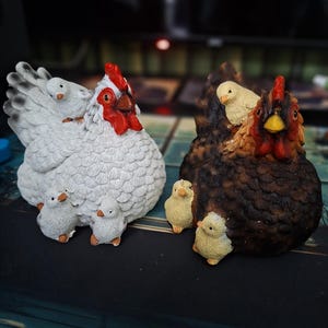 Puede incluir: Dos figuras decorativas de gallinas. Una es blanca con detalles negros y varios pollitos pequeños. La otra es marrón con detalles rojos y amarillos, también con pollitos. Ambas tienen detalles y probablemente están hechas de resina o material similar.