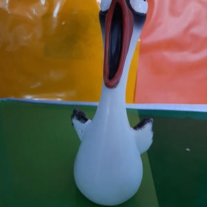 Könnte beinhalten: Eine weiße Keramikfigur eines Vogels mit langem Hals und offenem Schnabel. Der Vogel hat große, runde Augen und schwarze Markierungen an seinen Flügeln. Die Figur steht vor einem bunten Hintergrund.