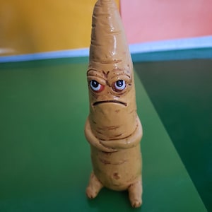 Puede incluir: Una figurita de cerámica amarilla en forma de zanahoria con una expresión de enfado. La zanahoria tiene los brazos cruzados y está de pie sobre una superficie verde.