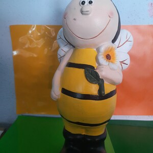 Puede incluir: Una figura de abeja sonriente con un cuerpo a rayas amarillas y negras, botas negras y alas blancas. La abeja sostiene una flor blanca con un centro naranja. La figura tiene antenas negras con puntas amarillas.